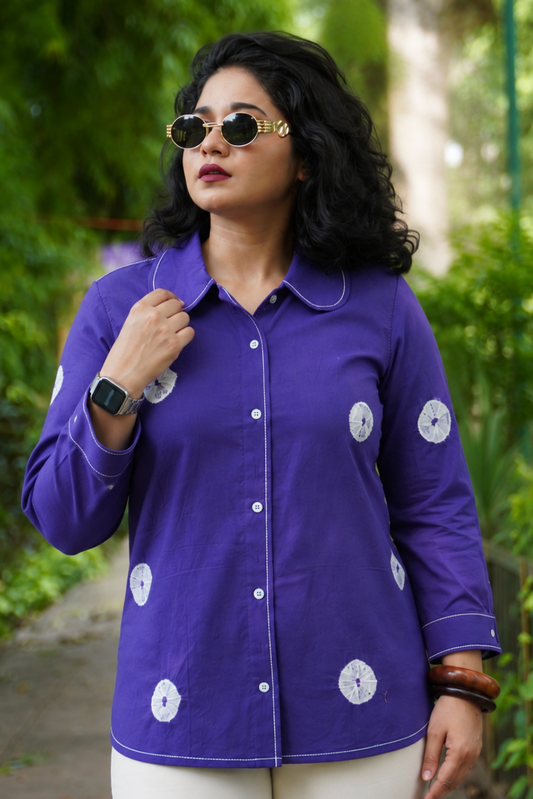 A woman wearing a purple long-sleeve button-up shirt with white circular patterns, styled with white pants and gold-framed sunglasses, standing outdoors on a garden path.