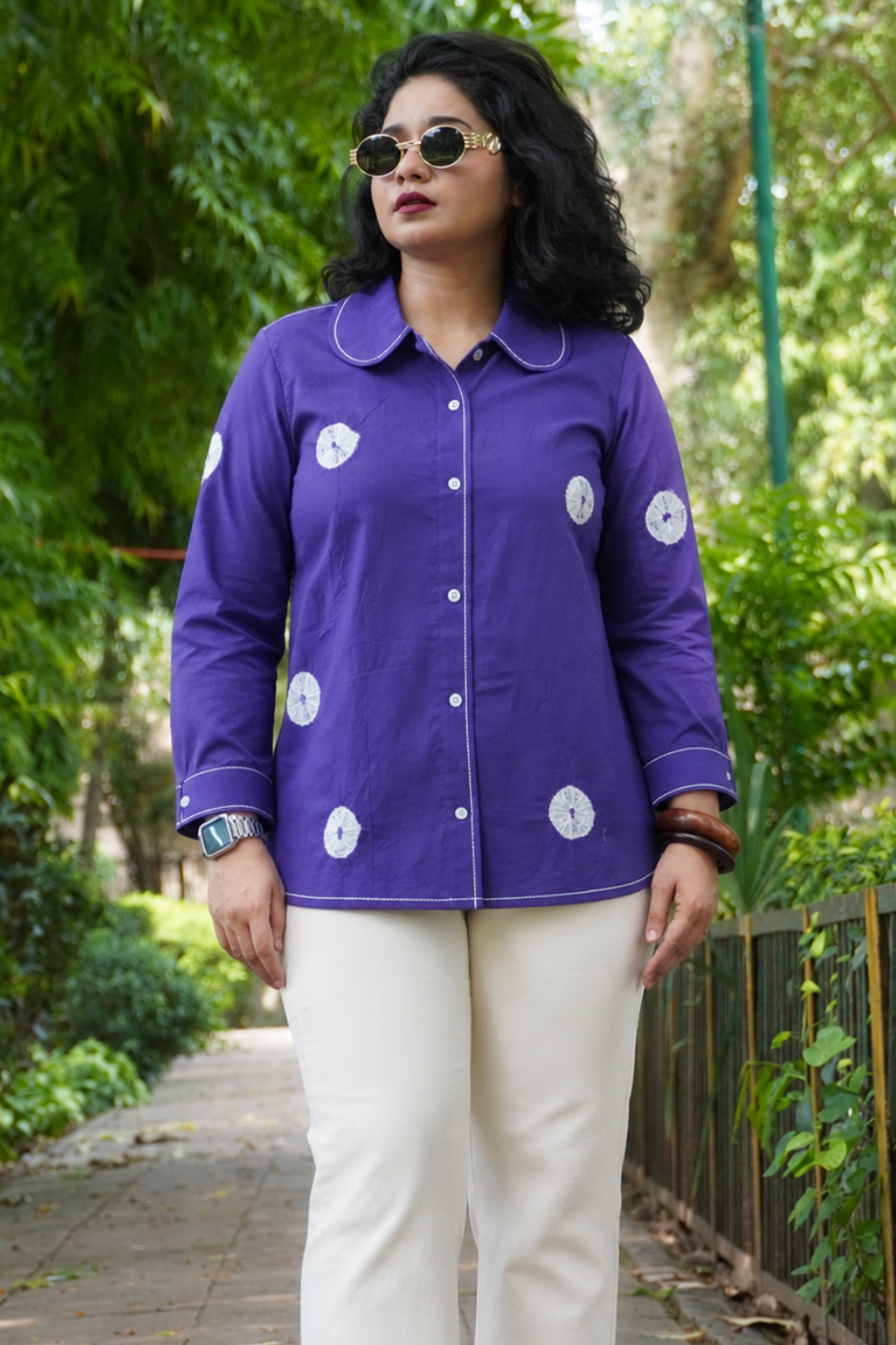 A woman wearing a purple long-sleeve button-up shirt with white circular embroidered motifs and cream pants, standing on a paved path in a lush green outdoor setting.